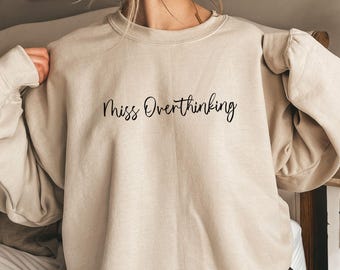 May include: Sand-colored sweatshirt with the text "Miss Overthinking" in a black script. The long-sleeved top is made of a soft material. The sweatshirt is a casual style.