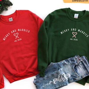 May include: A red sweatshirt and a forest green sweatshirt, each featuring the white text MERRY AND MARRIED, a graphic of two crossed candy canes forming a heart, and the text est. 2026. A pair of blue denim jeans is placed over the green sweatshirt.