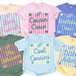 May include: Six short-sleeve t-shirts in ivory, blossom, flo blue, light green, chambray, and butter colors. Each shirt features a central graphic with vertical stripes, a paperclip icon, star accents, and text including Best Friends, Cousin Crew, Birthday Crew, The Johnsons, Cool Cousins, and Little Artist.
