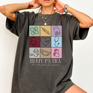 May include: A dark gray oversized t-shirt with a graphic print of medical icons in a grid. The text "IN MY PA ERA" and "Michelle's Version" is printed below the graphic.