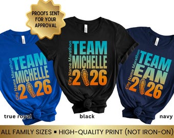 Puede incluir: Tres camisetas de manga corta en azul real, negro y azul marino. Cada camiseta presenta un gráfico con el texto "TEAM MICHELLE 2026" o "TEAM EAN 2026" y "City Name Marathon" con un icono de zapatilla de correr en el centro.