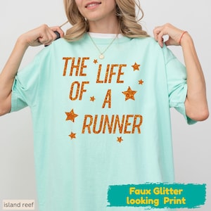 May include: A light mint green short-sleeve t-shirt featuring the orange glitter-effect text THE LIFE OF A RUNNER arranged in three lines, accented by several orange glitter-effect stars scattered around the lettering on the front.