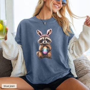 May include: A blue jean t-shirt with a graphic of a raccoon wearing pink bunny ears and holding a colorful Easter egg.