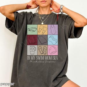 May include: A charcoal gray t-shirt featuring a grid of nine illustrations related to swimming, including goggles, fins, a swimsuit, a towel, a kickboard, a pool noodle, a tumbler, flip-flops, and a swim cap, with the text "IN MY SWIM MOM ERA" and "Michelle's Version".