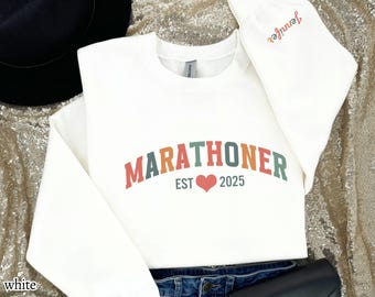 May include: White sweatshirt with a colorful "MARATHONER EST 2025" graphic with a red heart. The sweatshirt is laid out on a gold sparkly surface.