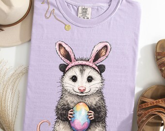 Può includere: T-shirt Comfort Colors color orchidea con un opossum con orecchie da coniglio rosa e un uovo di Pasqua colorato. La maglietta ha un girocollo e maniche corte. Una collana d'oro e un cappello beige sono visibili sullo sfondo.