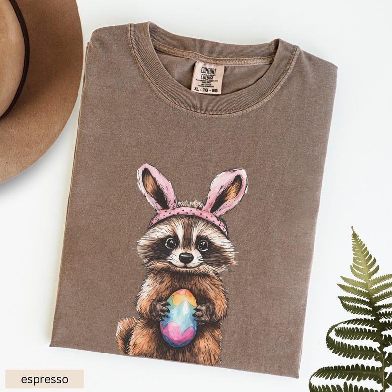 May include: A brown t-shirt with a graphic of a raccoon wearing pink bunny ears and holding a colorful Easter egg. The t-shirt is made of a soft, comfortable fabric and has a distressed look. The text "COMFORT COLORS" and "XL - T0 - 50" is printed on a white tag attached to the shirt.