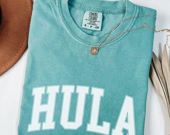 May include: Seafoam green t-shirt with the word "HULA" in bold white letters. The shirt has a crew neck and short sleeves. A gold necklace with a pendant is draped across the shirt. The word "seafoam" is printed at the bottom.