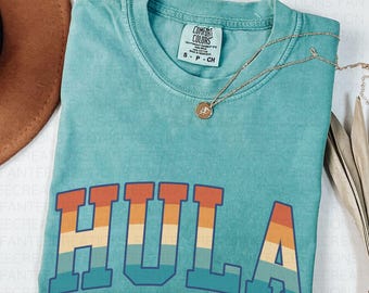 May include: Seafoam green t-shirt with the word "HULA" in a retro font. The letters are multi-colored with orange, yellow, and blue stripes. A gold necklace with a pendant is draped on the shirt. The shirt is labeled "Comfort Colors".