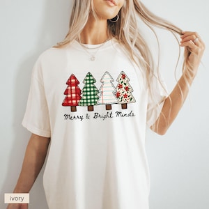 May include: Ivory-colored t-shirt featuring four Christmas tree designs with various patterns: red plaid, green gingham, lined paper, and floral. The text "Merry & Bright Minds" is printed below the trees. A casual, festive apparel item.