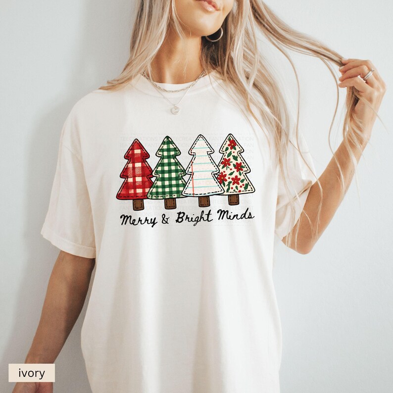 May include: Ivory-colored t-shirt featuring four Christmas tree designs with various patterns: red plaid, green gingham, lined paper, and floral. The text "Merry & Bright Minds" is printed below the trees. A casual, festive apparel item.