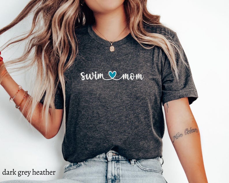 以下が含まれることがあります： 濃いグレーのヘザーTシャツに、白い文字で「swim mom」と、青いハートが書かれています。