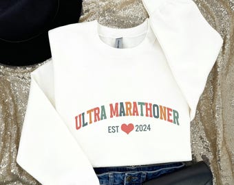 May include: Cream-colored sweatshirt with the text "ULTRA MARATHONER" in orange, green, and red lettering. Below the text is a red heart and the year "2024". The sweatshirt is folded on top of blue jeans.