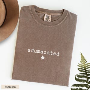 May include: A brown t-shirt with the text "edumacated" and a white star printed on the front. The shirt is made of a soft, comfortable material.
