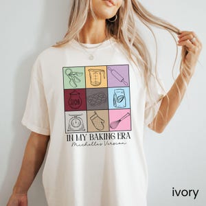 May include: A white t-shirt featuring a graphic of baking tools and the text "In My Baking Era Michelle's Version".