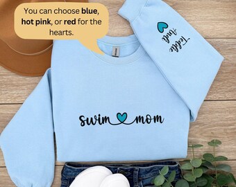 May include: A light blue crewneck sweatshirt featuring the black text "swim mom" with a teal heart in the center. The sleeve displays a teal heart above the black text "Andi Teddie". A speech bubble states: "You can choose blue, hot pink, or red for the hearts."