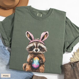 May include: A green t-shirt with a cartoon raccoon wearing pink bunny ears and holding a colorful Easter egg. The shirt has the text "Comfy Crew" printed on the tag.