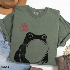 May include: A moss green t-shirt with a black and white graphic of a frog and a red stamp with Japanese characters. The shirt is made of a soft, comfortable material.