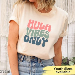 May include: Cream-colored t-shirt with the text "HULA VIBES ONLY" in pink, green, and blue retro font. The shirt is paired with blue jeans. The words "Youth Sizes available" are also visible.
