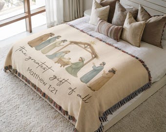 May include: A cream-colored woven throw blanket featuring a nativity scene illustration and the text "the greatest gift of all Matthew 1:21". The blanket has a fringed edge and depicts figures gathered around a manger under a star.