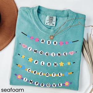 May include: Seafoam green t-shirt with a graphic of colorful beaded garlands spelling out the words "ASK IMAGINE PLAN CREATE IMPROVE".