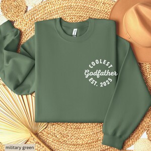 May include: A military green sweatshirt with the text "Coolest Godfather Est. 2025" in white.