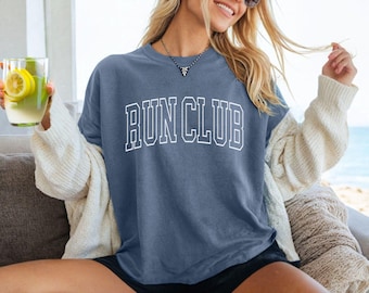 May include: A woman wearing a blue jean shirt with the text "RUN CLUB" in white outlined letters. She is wearing a white cardigan and black shorts.