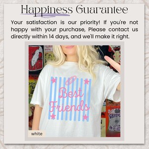 May include: A white t-shirt featuring a graphic design with vertical light blue stripes, pink stars at the corners, and the text "Best Friends" in pink script. The top of the graphic includes a pink paperclip icon. The shirt is white.