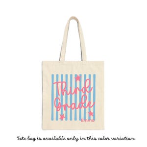 May include: A beige canvas tote bag featuring a vertical light blue striped pattern. The text "Third Grade" is printed in a pink script font, accompanied by three small pink stars and the name "Emma" in a simple pink sans-serif font.