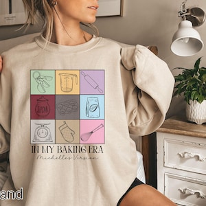 May include: A beige sweatshirt with a graphic design of baking tools and the text "In My Baking Era Michelle's Version".