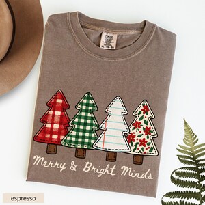May include: Espresso-colored t-shirt featuring four decorative Christmas trees with various patterns: red plaid, green gingham, lined paper, and floral. The text "Merry & Bright Minds" is printed below the trees.