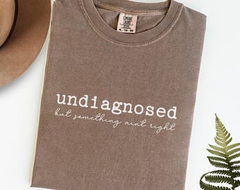 以下が含まれることがあります： エスプレッソブラウンのTシャツに、白い文字で「undignosed but something ain't right」と書いてあります。