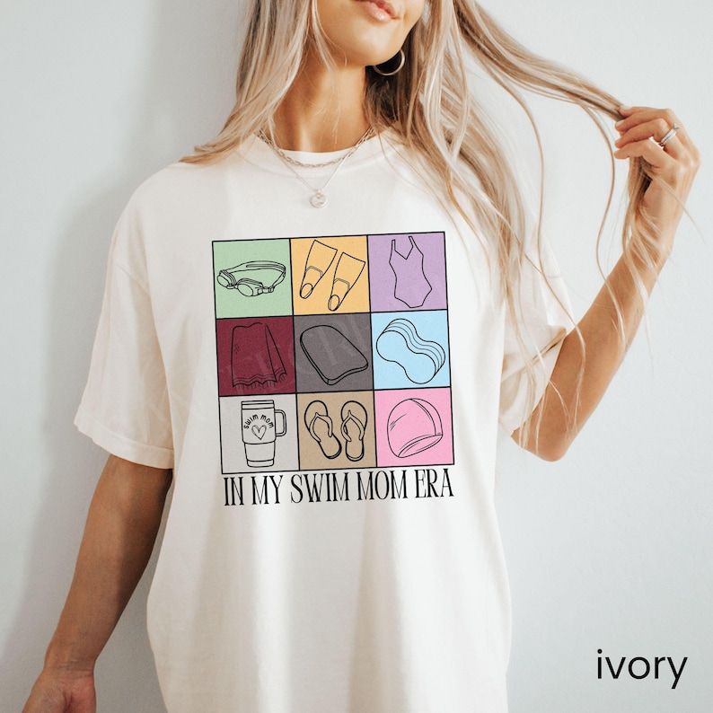 May include: An ivory t-shirt featuring a grid of nine colorful squares, each containing a black line drawing of swimming gear including goggles, fins, a swimsuit, a towel, a kickboard, a pool noodle, a tumbler, flip-flops, and a cap, with the text IN MY SWIM MOM ERA.