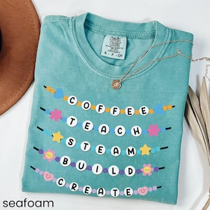 May include: Seafoam green t-shirt with a graphic of a colorful beaded garland spelling out the words "Coffee, Teach, Steam, Build, Create".