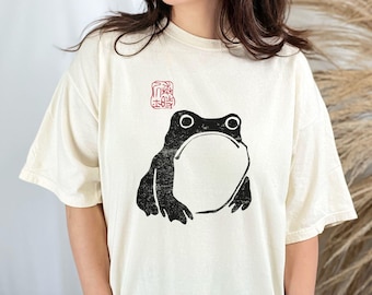 May include: A cream-colored t-shirt with a black and white graphic of a frog. The frog is in a simple, minimalist style and has a red stamp above it that says "lucky frog".