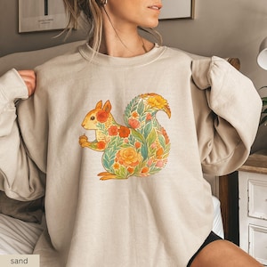 May include: Sand-colored sweatshirt featuring a detailed illustration of a squirrel. The squirrel is adorned with a floral pattern in shades of orange, red, and green. The sweatshirt has a crew neck and long sleeves, perfect for casual wear.