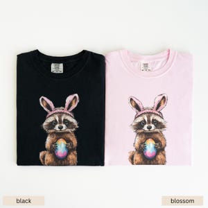 May include: Two t-shirts featuring a raccoon wearing bunny ears and holding a colorful Easter egg. The t-shirts are available in black and pink.