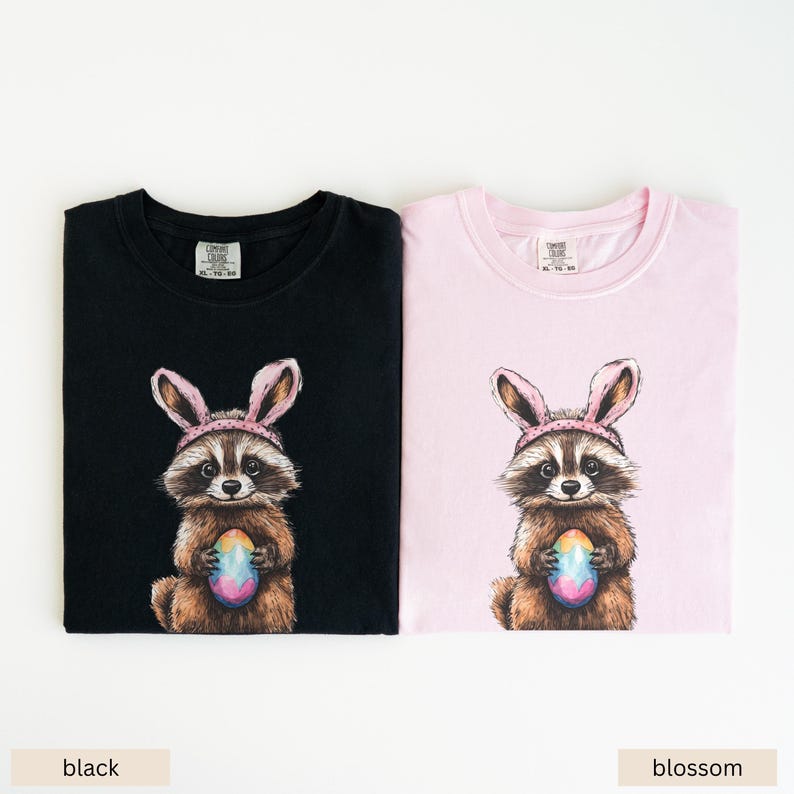 May include: Two t-shirts featuring a raccoon wearing bunny ears and holding a colorful Easter egg. The t-shirts are available in black and pink.