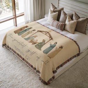 May include: A beige throw blanket featuring a watercolor illustration of the Nativity scene. The blanket has a fringed edge with a multi-colored pattern. Text on the blanket reads "the greatest gift of all" and "Matthew 1:21".