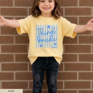 May include: A butter yellow short-sleeved t-shirt featuring a graphic design with vertical light blue stripes, blue stars, and the blue text "Third Grade" with the name "Abby" printed below in a smaller blue font on the front center.