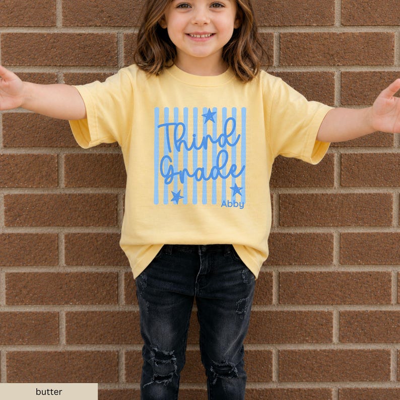 May include: A butter yellow short-sleeved t-shirt featuring a graphic design with vertical light blue stripes, blue stars, and the blue text "Third Grade" with the name "Abby" printed below in a smaller blue font on the front center.