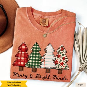 May include: A coral-colored t-shirt with a graphic of four Christmas trees in red plaid, green gingham, white with blue lines, and red flowers. The text "Merry & Bright Minds" is below the trees. A gold necklace and a brown hat are also visible.