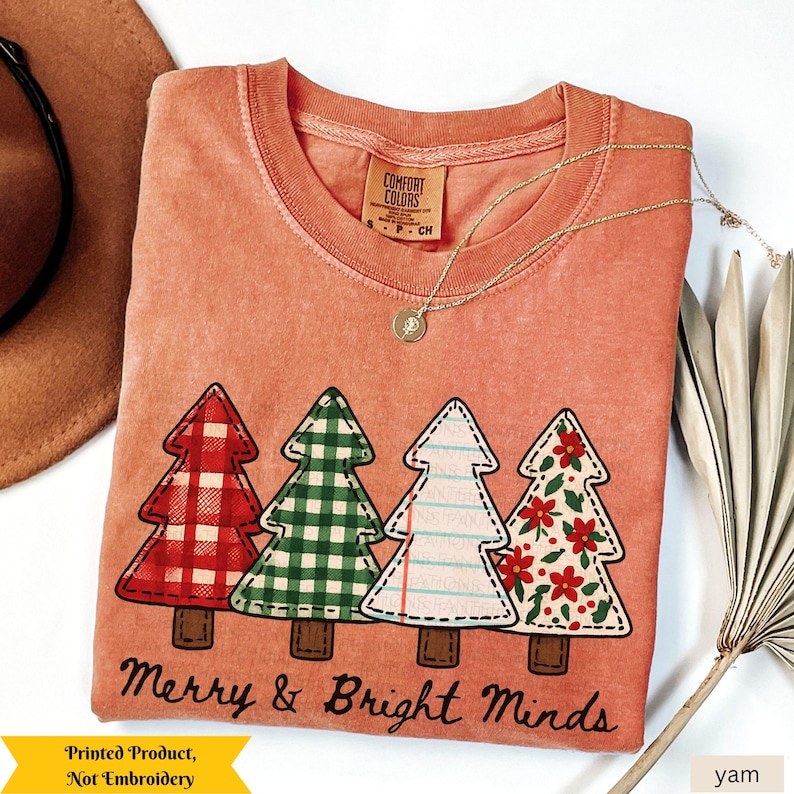 May include: A coral-colored t-shirt with a graphic of four Christmas trees in red plaid, green gingham, white with blue lines, and red flowers. The text "Merry & Bright Minds" is below the trees. A gold necklace and a brown hat are also visible.