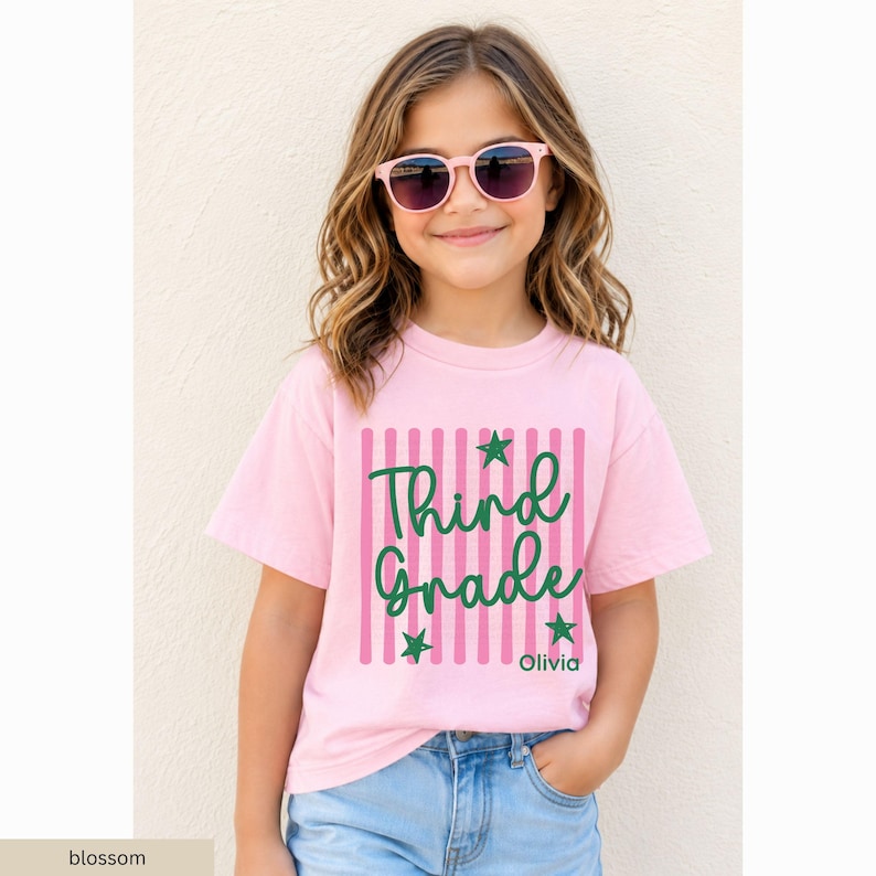 May include: A light pink short-sleeved t-shirt featuring a vertical striped pattern in a darker pink shade. The center graphic displays the dark green text "Third Grade" in a cursive font, accented by three small green stars and the name "Olivia".