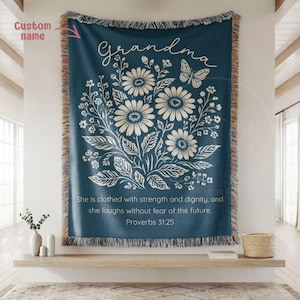 May include: A navy blue woven throw blanket with a floral design and the word "Grandma" in script. The blanket features a butterfly and the Bible verse Proverbs 31:25. The edges have a fringed trim.