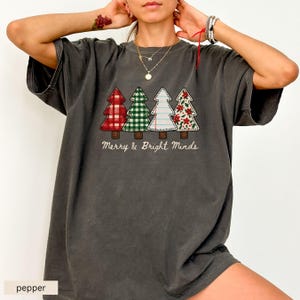 May include: A dark gray t-shirt featuring four Christmas trees with different patterns: red plaid, green gingham, white with stitching, and a floral design. The text "Merry & Bright Minds" is below the trees.