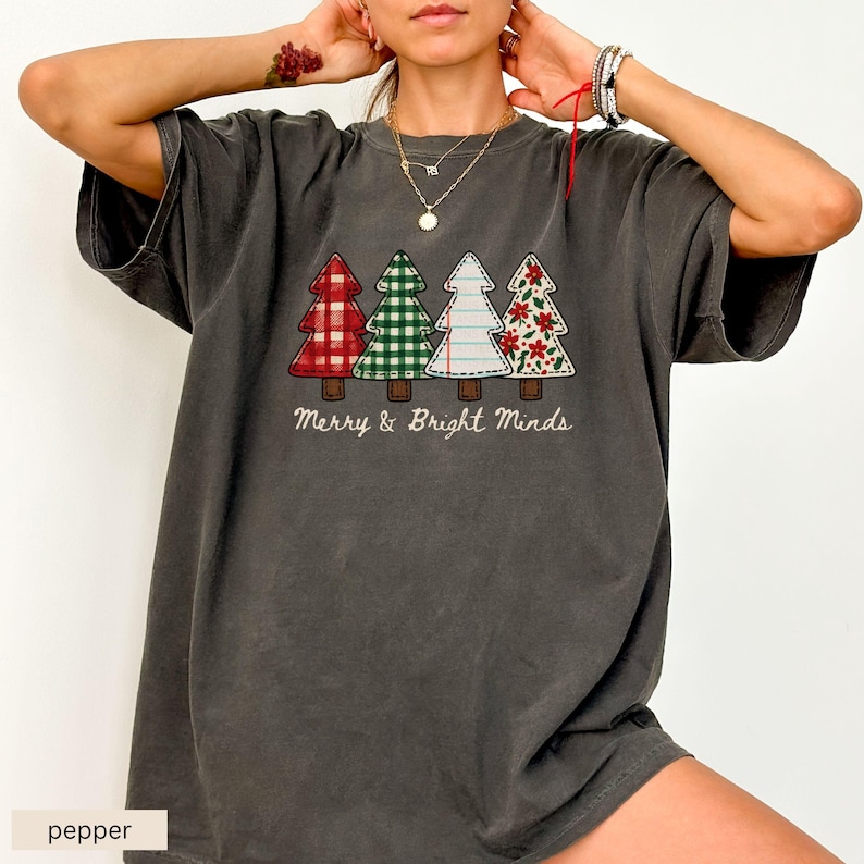 May include: A dark gray t-shirt featuring four Christmas trees with different patterns: red plaid, green gingham, white with stitching, and a floral design. The text "Merry & Bright Minds" is below the trees.