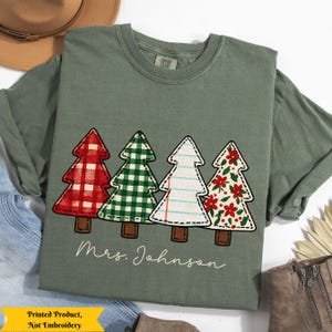 May include: Sage green long-sleeve t-shirt featuring four Christmas tree designs. The trees have red plaid, green gingham, lined paper, and floral patterns. The name "Mrs. Johnson" is printed below the trees.