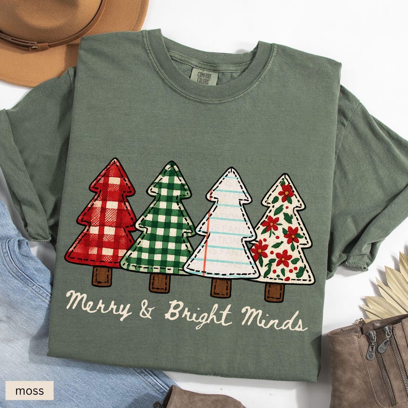 May include: A moss green t-shirt featuring four Christmas tree designs with different patterns and colors. The trees are red plaid, green gingham, lined paper, and floral. The text "Merry & Bright Minds" is printed below the trees.