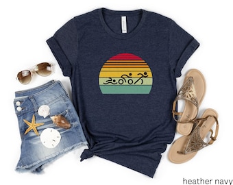 May include: Heather navy blue t-shirt featuring a vintage-style sunset graphic with a silhouette of a group of swimmers. The graphic is in shades of yellow, orange, red, and green.